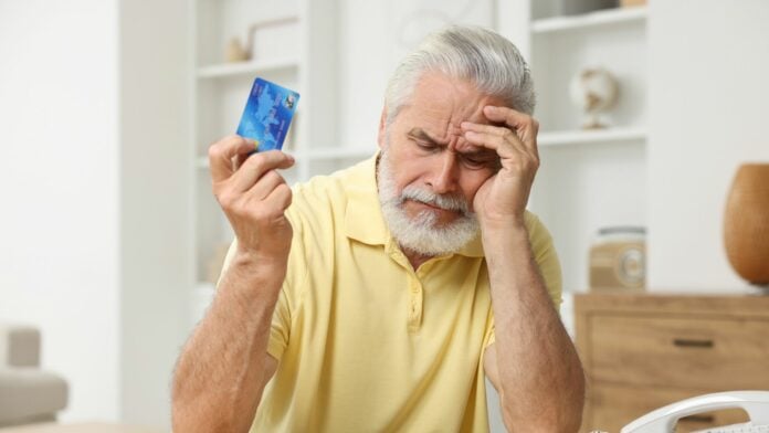 Idoso de expressão preocupada sentado em casa, segurando um cartão de crédito em uma mão e apoiando a outra na testa, representando tensão financeira.